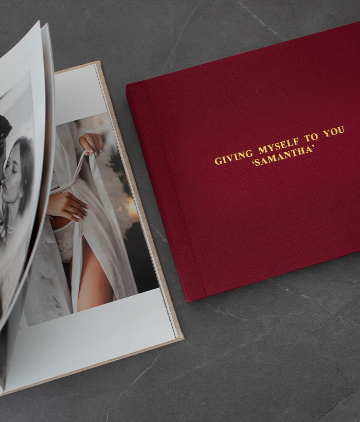 A red photo album with gold-embossed text lies beside an open album displaying prints of intimate images, with a layflat design highlighting the precise photo book printing and craftsmanship available from the online photo book maker, FlipChap.