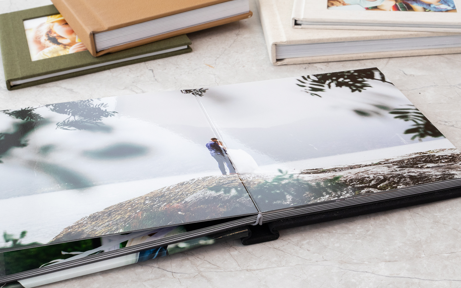 open wedding photobook on a marble surface showing a couple on a cliff surrounded by lush greenery with various photo albums in the background