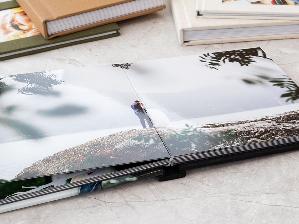 open wedding photobook on a marble surface showing a couple on a cliff surrounded by lush greenery with various photo albums in the background